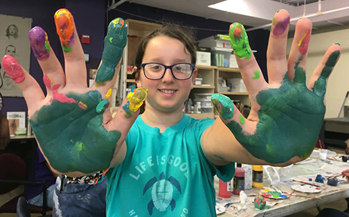 A young person in an art studio smiles and holds up their paint-covered hands