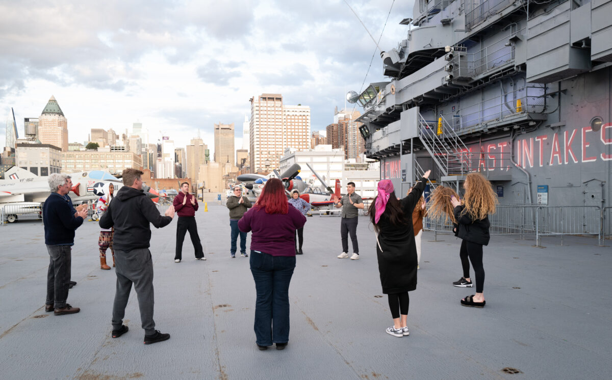 A group of people stand in a circle being led through a dance exercise on the deck of the Intrepid, with military planes and the Manhattan skyline behind them