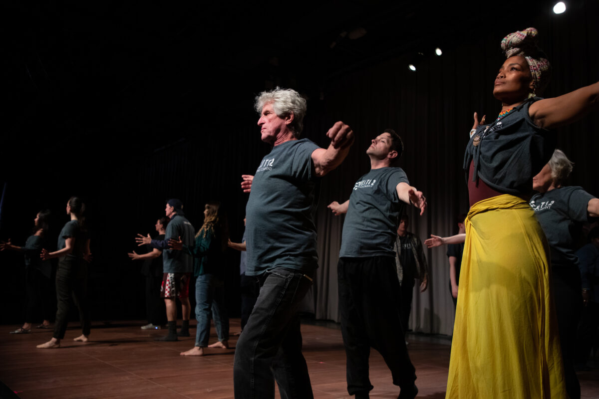 A group of dancers on stage with arms outstretched. An older white man with white hair is in the center of the frame, flanked by a younger white man and a woman of color in a headscarf and bright yellow skirt.