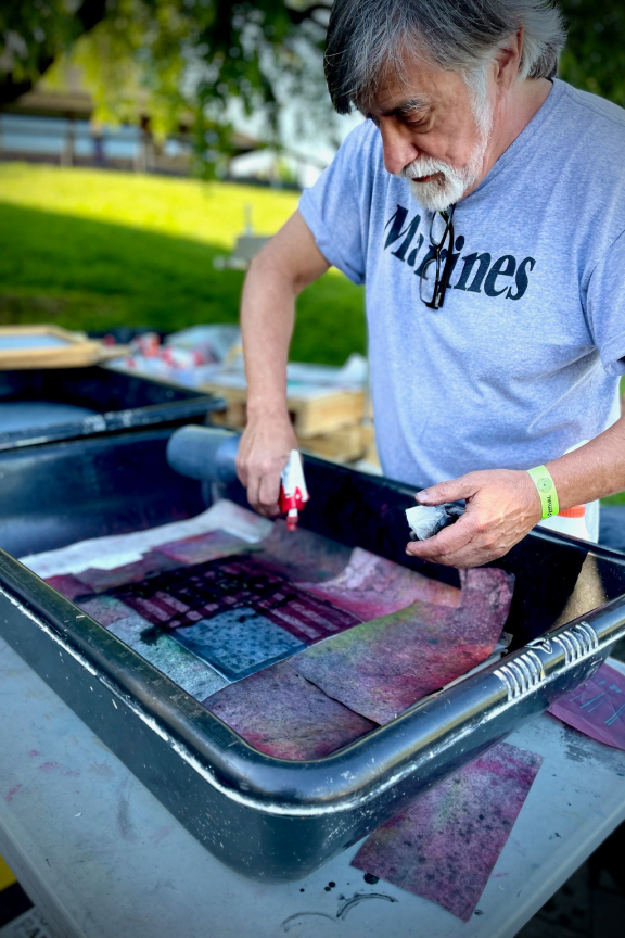 Man wearing a t-shirt that says "Marines" stands at a tub of homemade paper while holding a spray bottle in his right hand and a rag in high left hand.