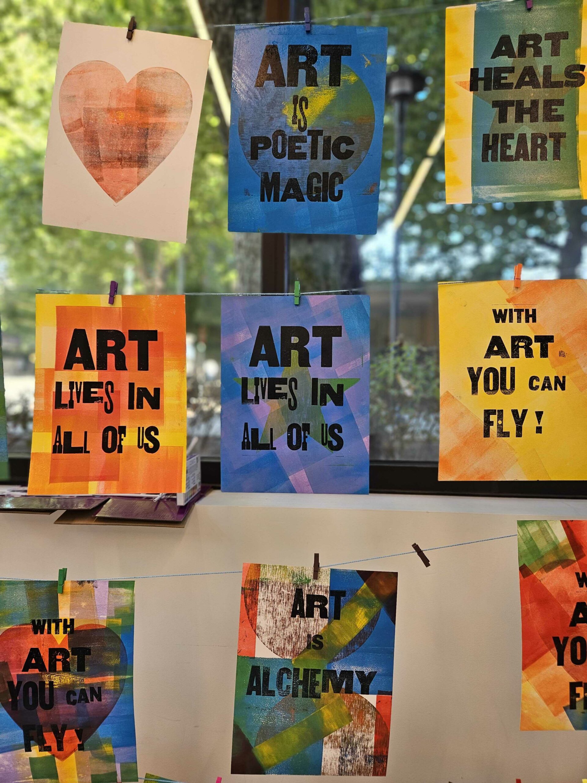 A wall of colorful prints hanging on strings with clothespins with messages reading "Art is Poetic Magic," "Art heals the heart," and "Art lives in all of us"