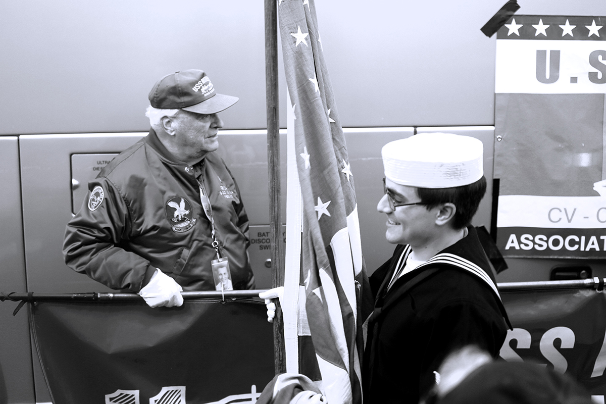 Black and white image of a side view of a man in a Navy uniform holding a flag while an older veteran look off to the right
