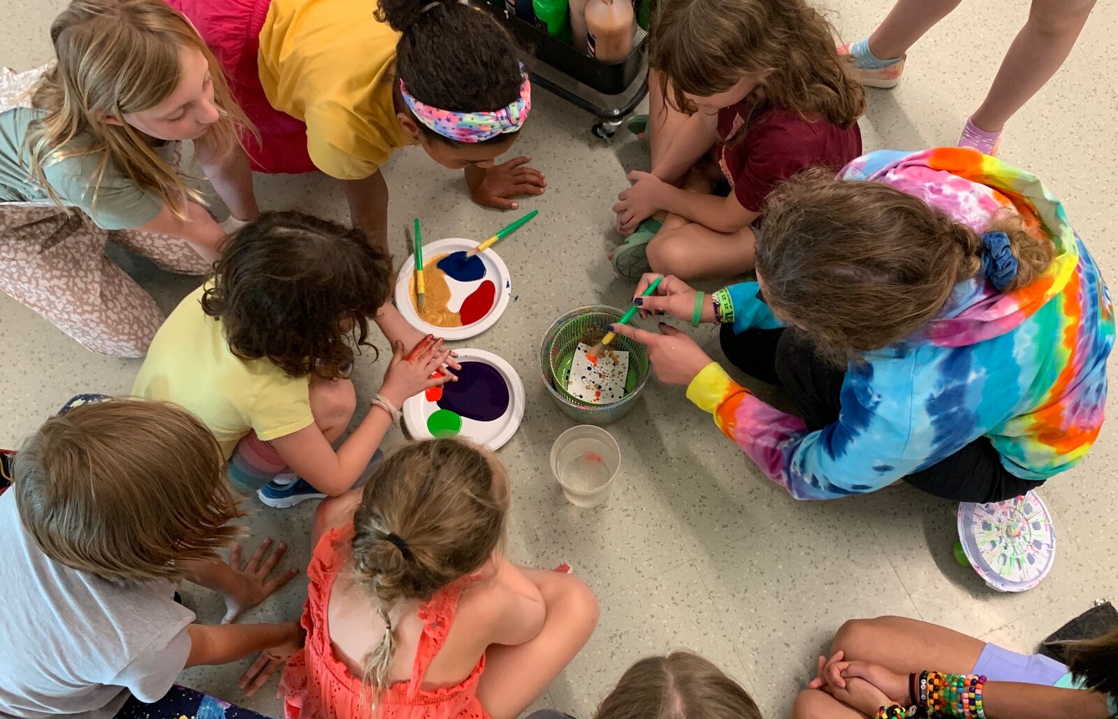 A group of children gather with a facilitator in a circle with paint and brushes at the center.