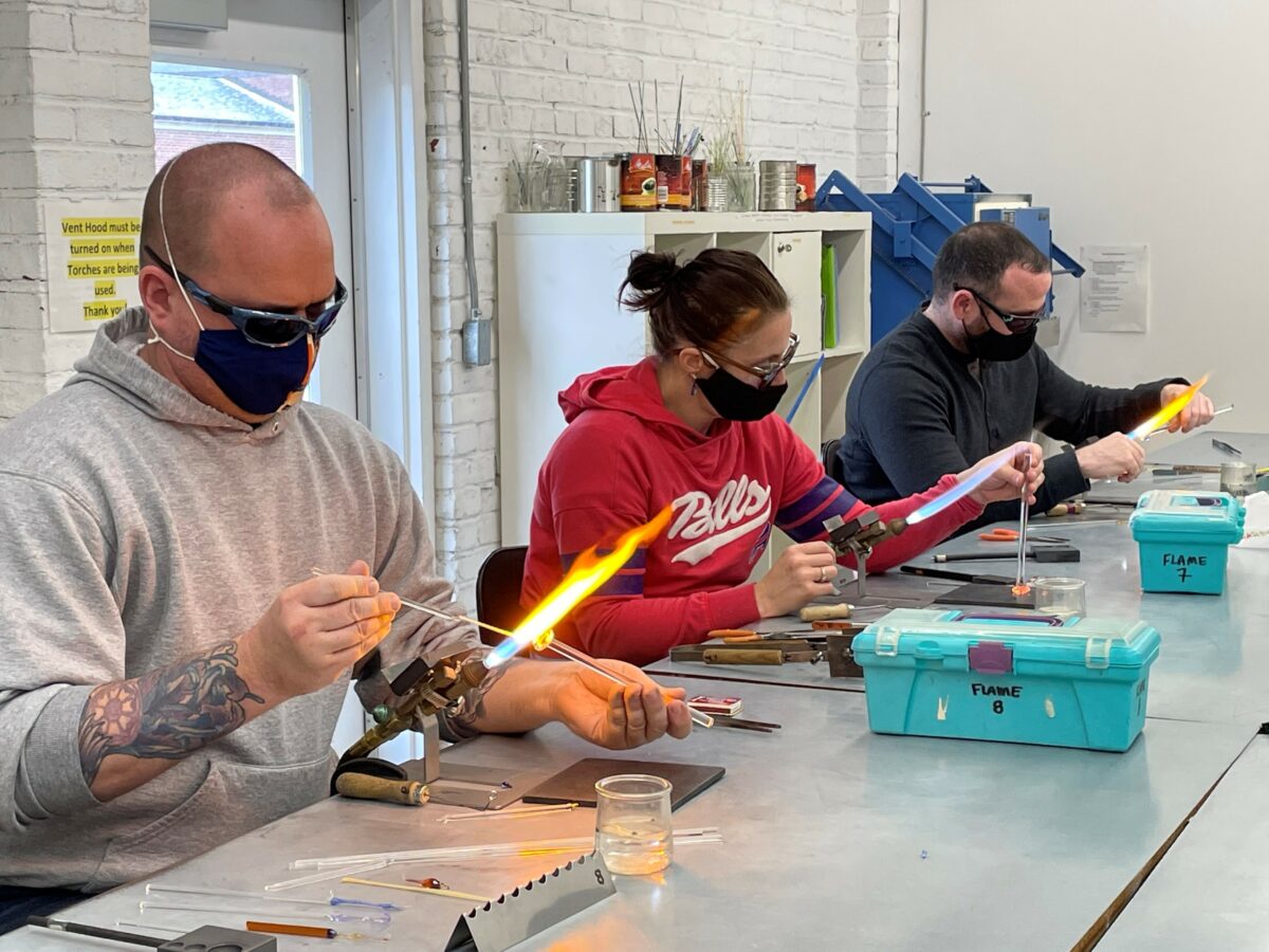 Three people seated at a table, each wearing protective eyewear, masks, and holding materials at a flame in a glass flameworking class.
