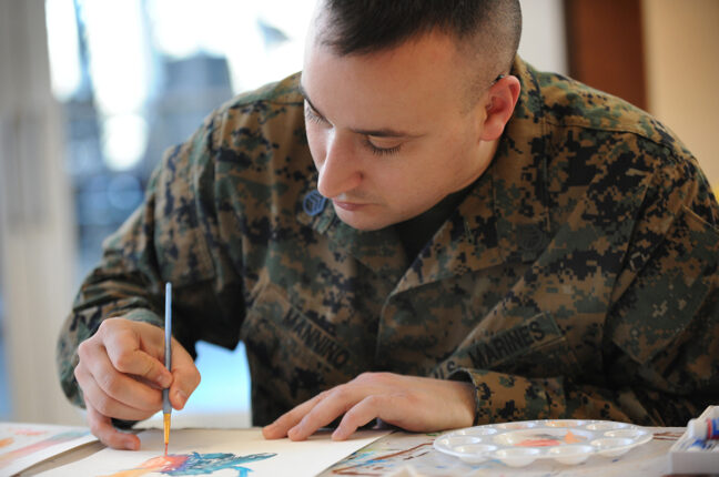 A white male with short brown hair wearing a Marines military uniform seated while holding a paintbrush and painting.