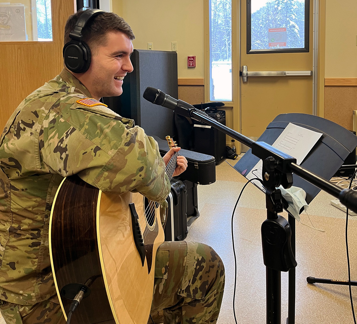 A white man with short dark hair wearing an Army uniform and headphones seated and smiling while holding a guitar. There is a microphone and music stand in front of him.
