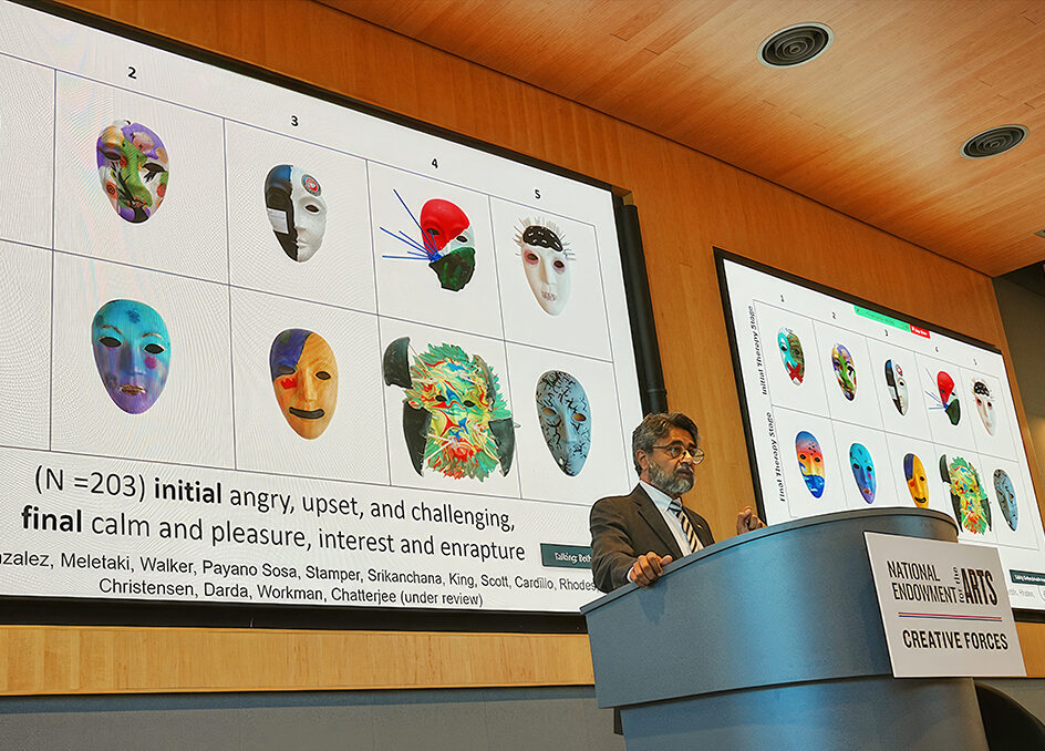 An Indian man with short black and grey hair and a black and grey beard wearing glasses while standing behind a podium. There is a screen behind him with images of decorated, colorful masks.
