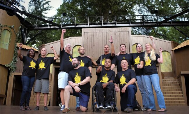 A group of performers on an outdoor stage. Eight people are standing with their hands or first in the air while two men kneel beside a performer in a wheelchair.