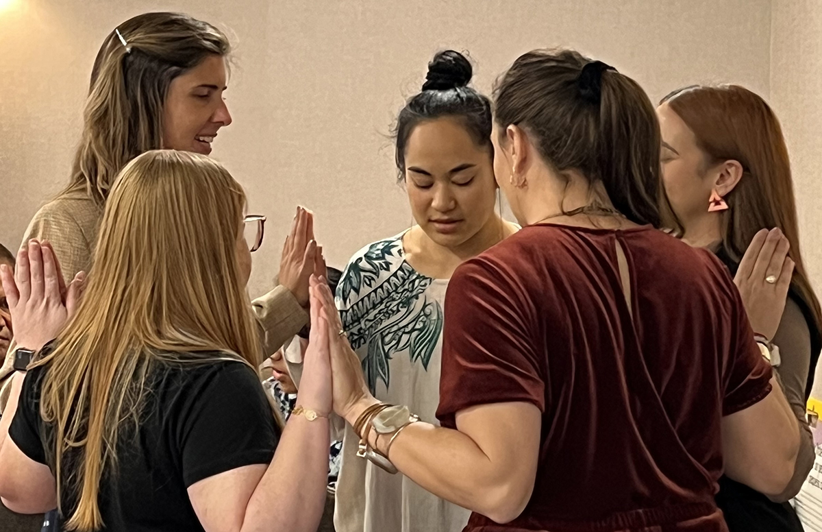 A group of women standing in a circle facing each other with each hand pressed together with the person on either side's hand.