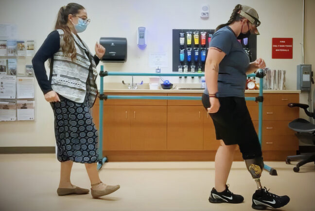 A dance therapist and her patient are shown walking in profile in a clinic. The patient has an artificial leg and is wearing black shorts.