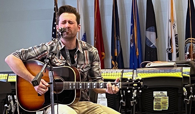 Man with short brown hair and his eyes closed seating while holding a guitar and singing into a microphone. There are flags on poles behind him.
