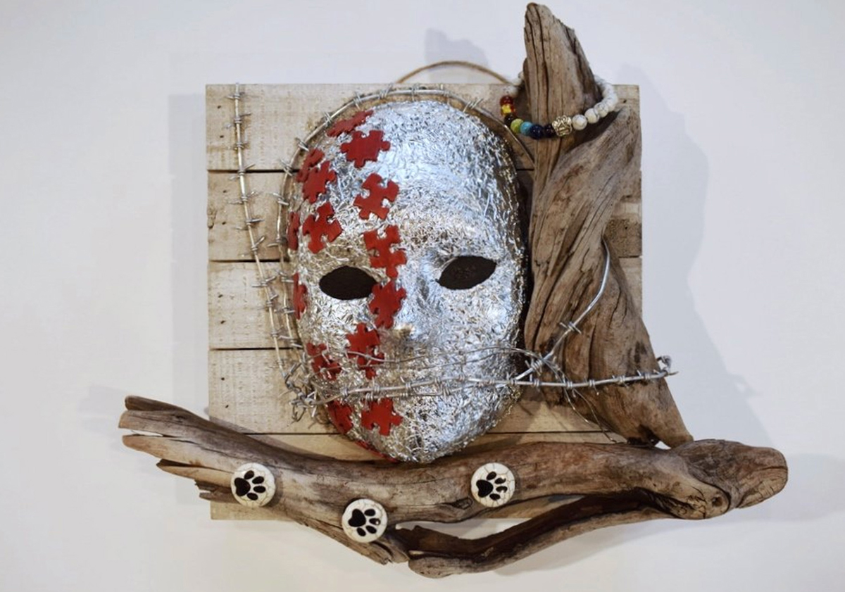 Mixed media sculpture with a silver mask with red puzzle pieces on it and barbed wire across it. The mask is mounted on wood and has wood branches on two sides. One branch has a bracelet and the other has animal prints.