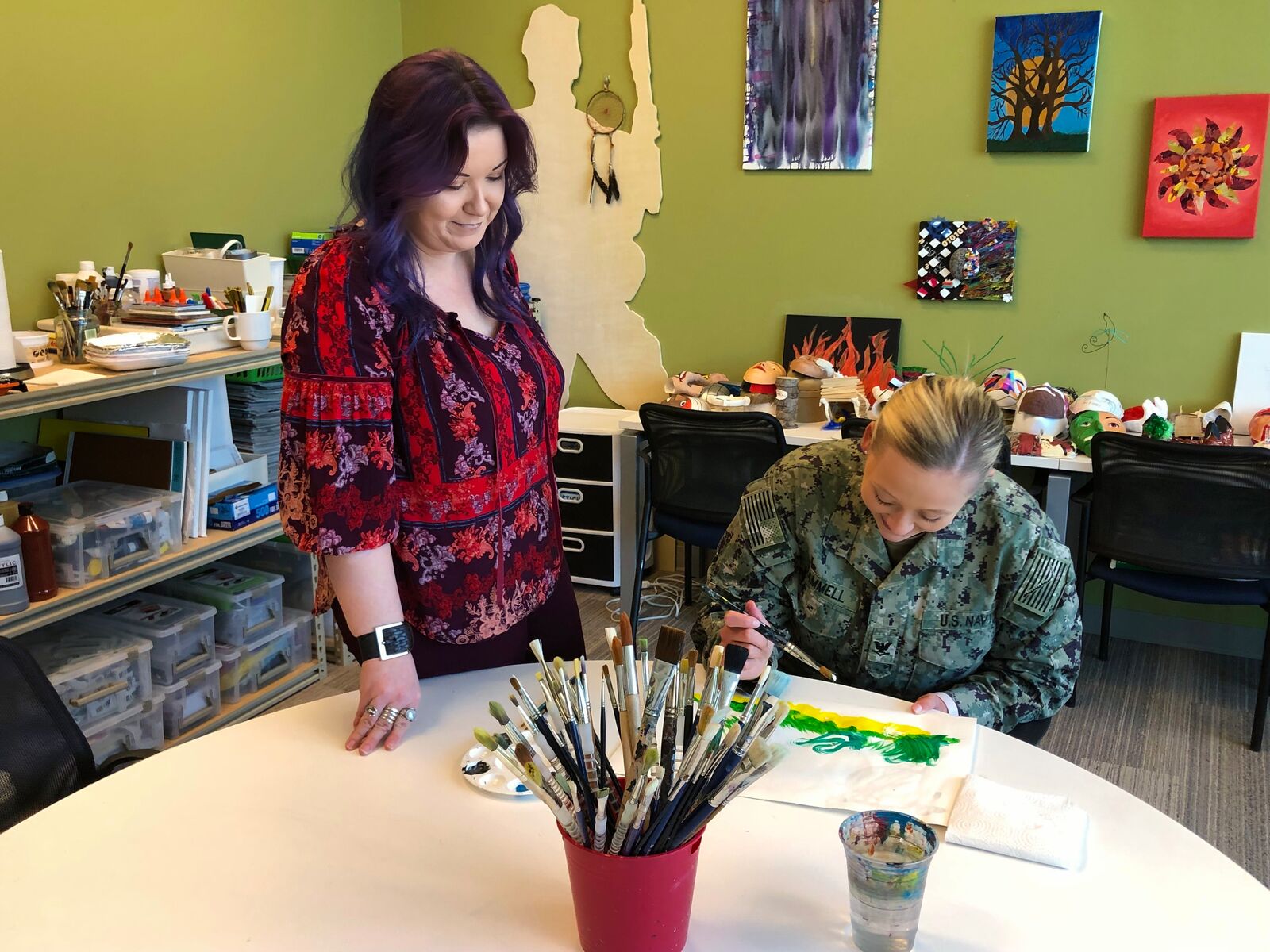 A white woman with long dark hair wearing a multi-colored red floral shirt is standing and looking down at a white woman with blond hair who is seated and wearing a military uniform. The seated woman is looking down while holding a paintbrush and painting. There is art on the walls and art supplies in the background.