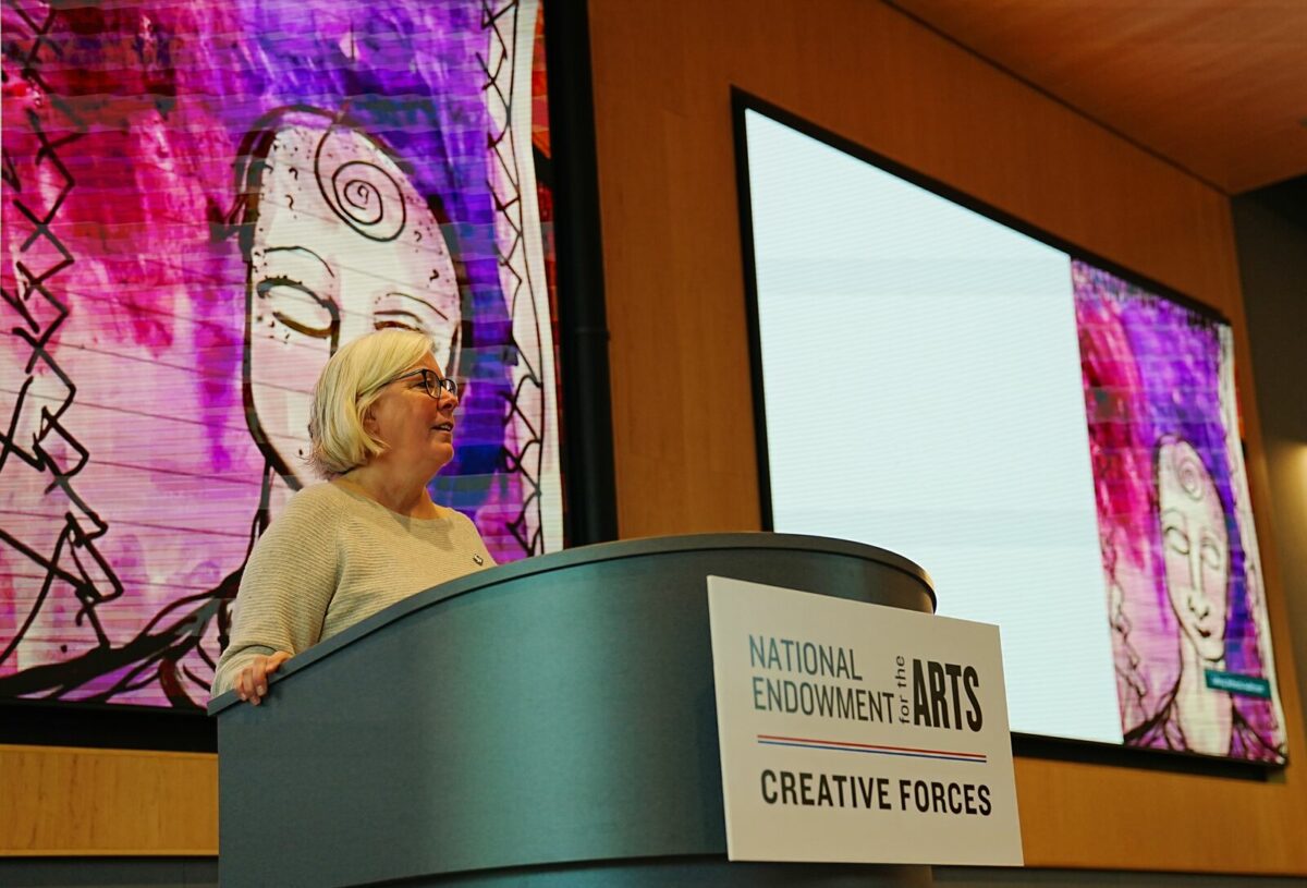 A white woman with white hair wearing glasses and a beige sweater standing behind a podium with a sign that says "National Endowment for the Arts Creative Forces." There is colorful artwork on the screens behind her.