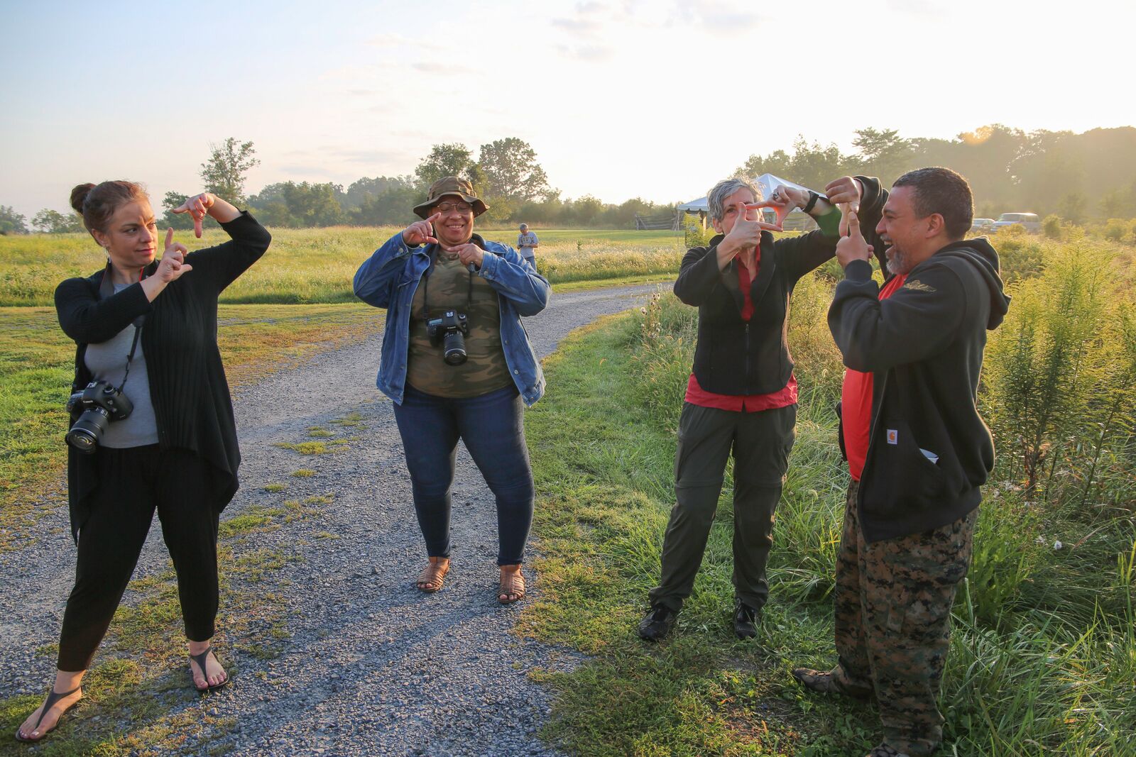 Four people standing outdoors on a sunny day with trees in the background and a path through grass. Two people have cameras that are hanging from their necks. Everyone is holding up their hands and looking through a square they made with their fingers.