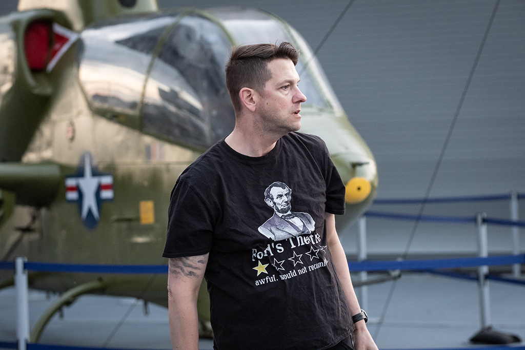 Image of a white male with short brown hair wearing a t-shirt with an image of President Lincoln while looking to the right. There is a historic military plane in the background.