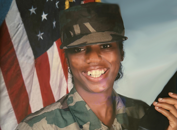 Portrait of a black woman in an Army uniform smiling while holding a gun. There is an American flag behind her.