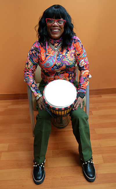 Photo of a black woman with shoulder-length hair smiling. She is wearing red glasses, a colorful shirt with flower patterns, and green pants while seated and holding a djembe drum.