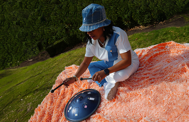 A black woman wearing a blue hat is seated on an orange furry blanket in a park.  She is holding mallets and playing a disc-shaped drum.