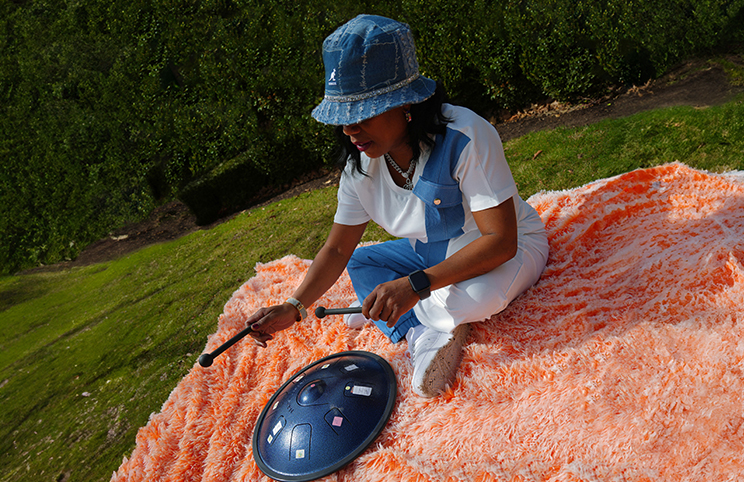A black woman wearing a blue hat is seated on an orange furry blanket in a park. She is holding mallets and playing a disc-shaped drum.