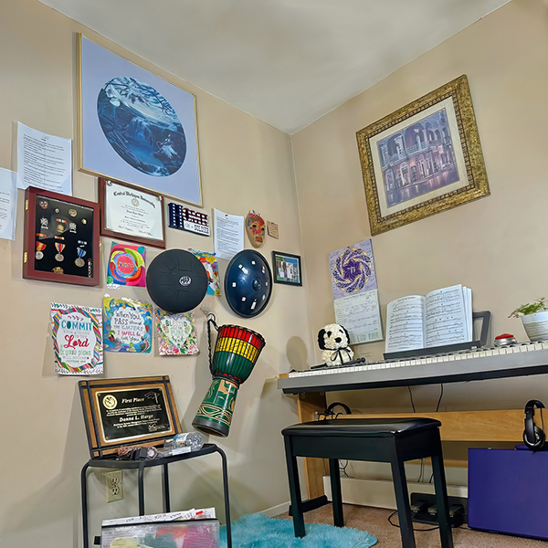 View of a corner of a room. There is art, military medals, a college degree, and percussion instruments mounted to one wall. Against the adjacent wall is a music keyboard with sheet music on top and a calendar and framed artwork on the wall above. There is a bench in front of the keyboard and headphones under the keyboard.