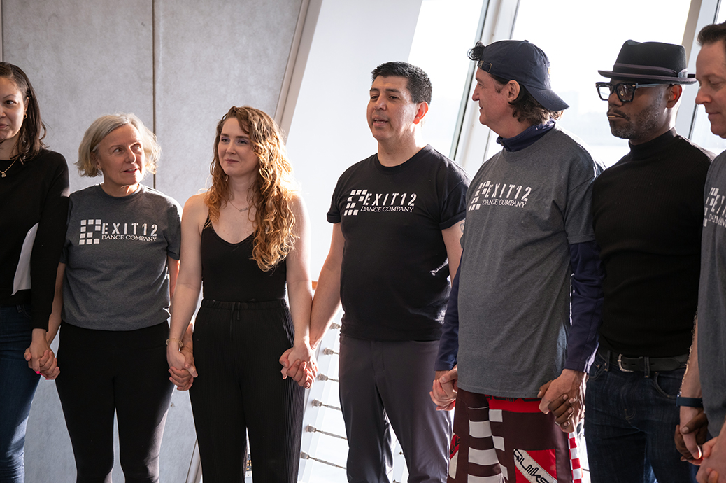 A man with dark hair wearing a black shirt that says "Exit 12 Dance Company" in white letters stands in the center of a group of male and female adults standing in a line and holding hands.