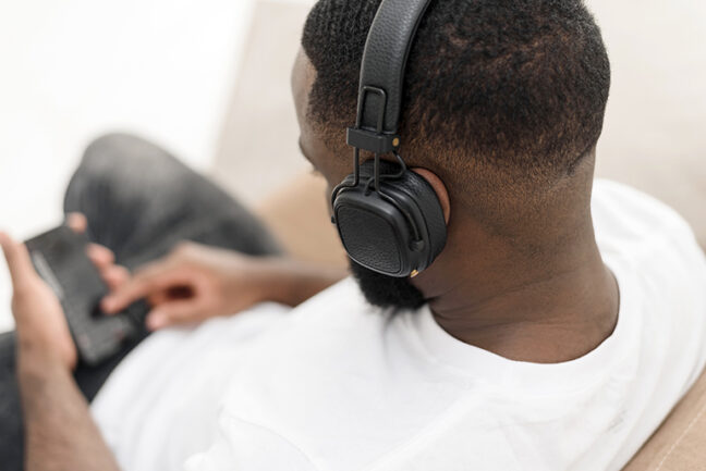 An over-the-shoulder view of a black male wearing headphones while looking down at a phone he is holding.