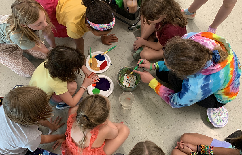 A group of children seated on the floor gathers with a facilitator in a circle with paint and brushes at the center.