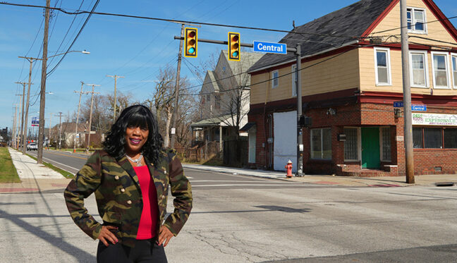 A black woman stands at a street intersection with her hands on her hips and smiling. The street sign behind her says "Central."