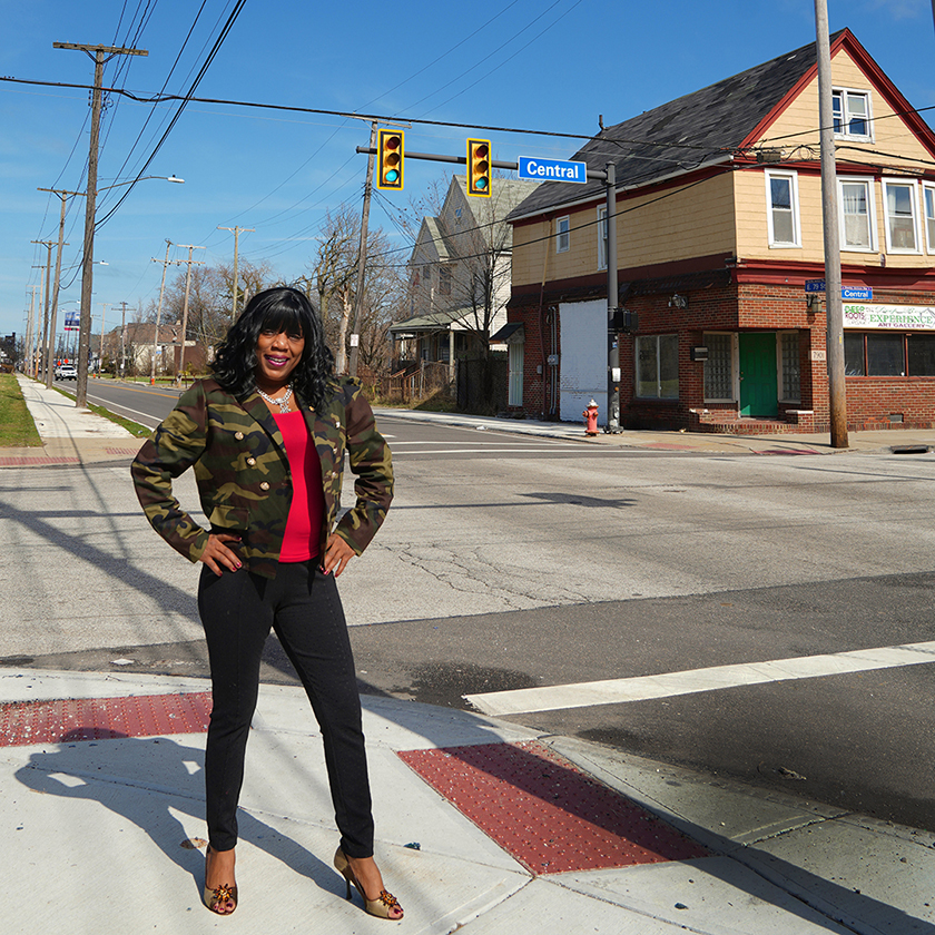 A black woman stands at a street intersection with her hands on her hips and smiling. The street sign behind her says "Central."