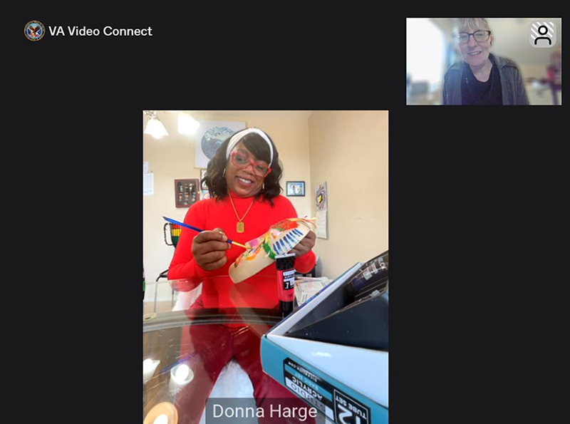 Screenshot of a virtual art therapy session. A black woman wearing glasses smiles as she paints a mask. A white woman with glasses smiles and watches at the top of the frame. The text at the top says VA Video Connect.