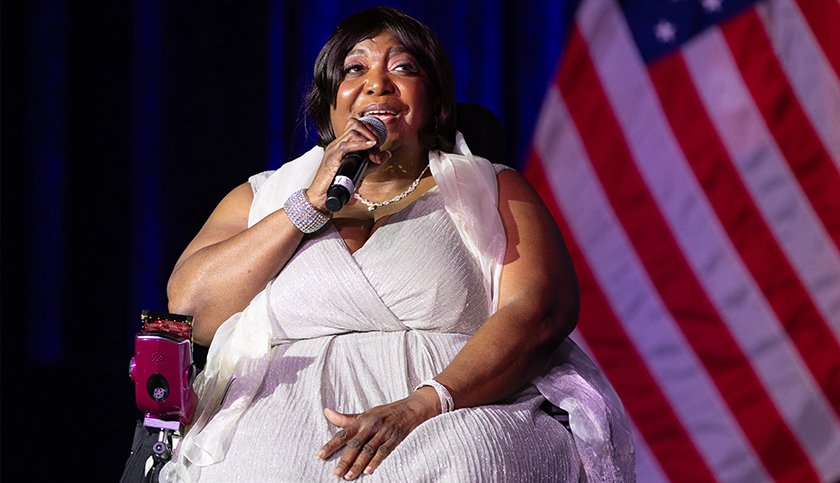 A black woman wearing a white sleeveless gown is seated and holding a microphone while singing. A blue stage curtain and an American flag are in the background.