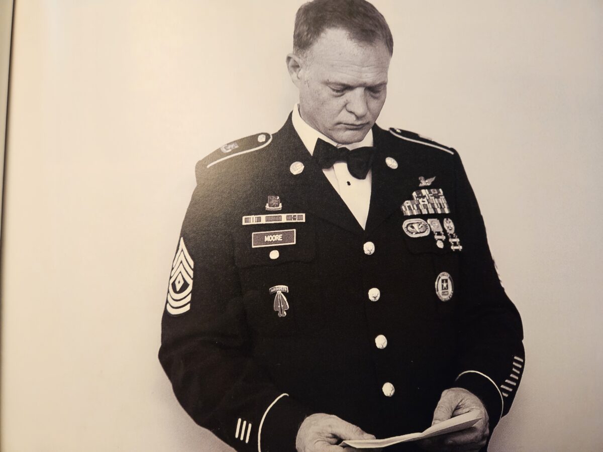 A man in a formal military uniform with various badges and insignia looks down at a piece of paper. The uniformed jacket features a name tag, and several medals and decorations are visible. The setting is simple and monochromatic.