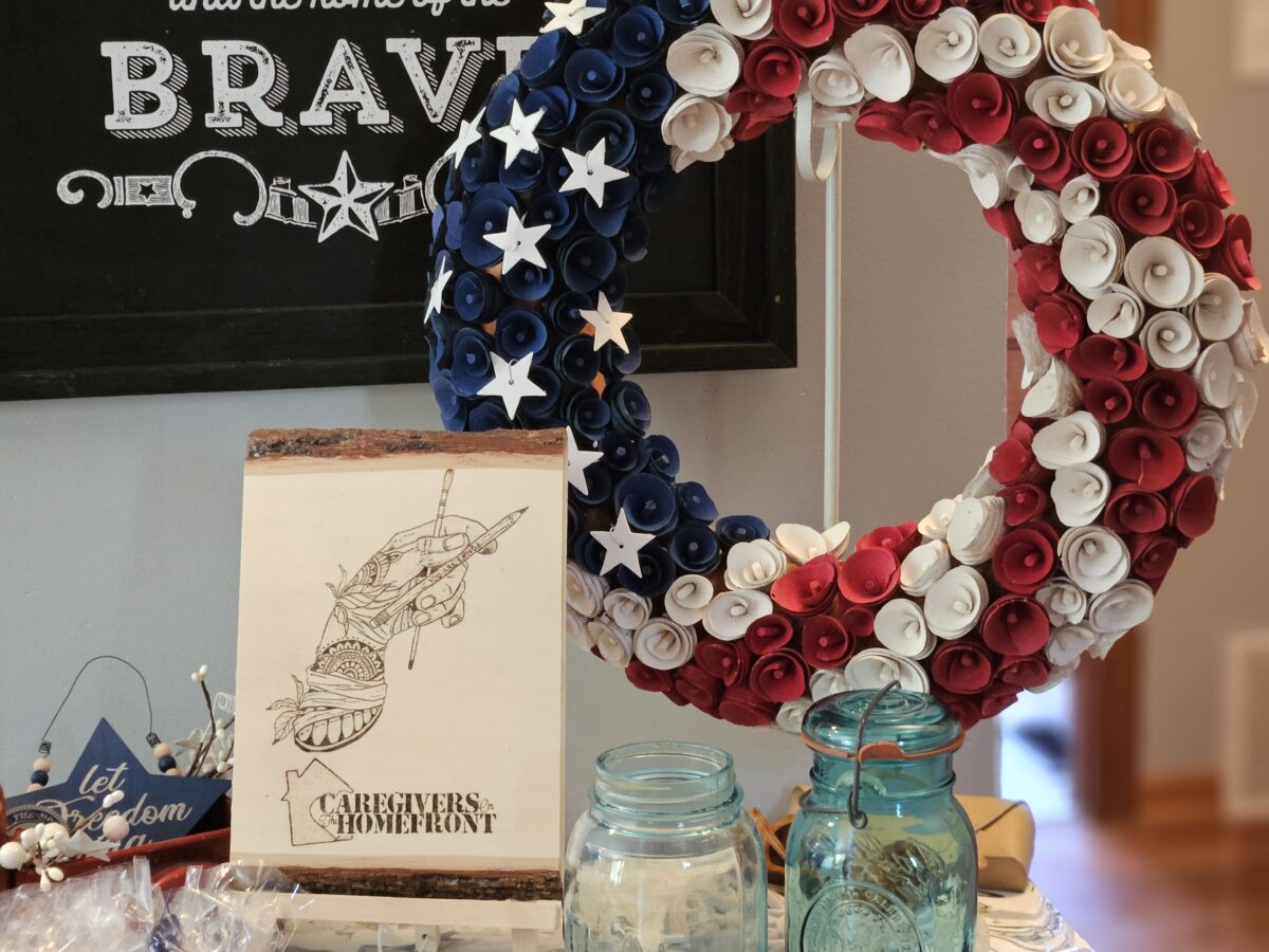A decorative wreath made of red, white, and blue paper flowers with stars hangs above a table. Below, two blue glass jars, a sign reading Let Freedom Ring, and a drawing titled Caregivers on the Homefront are displayed.