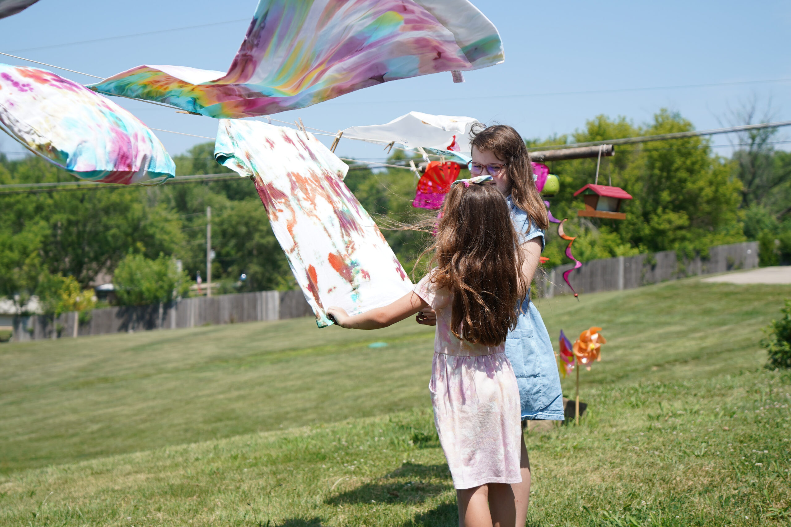 Two children hanging colorful tie-dyed shirts on a clothesline outside. One child in a blue dress, the other in a pink dress, both with long hair. They are in a grassy yard with trees and a house in the background.