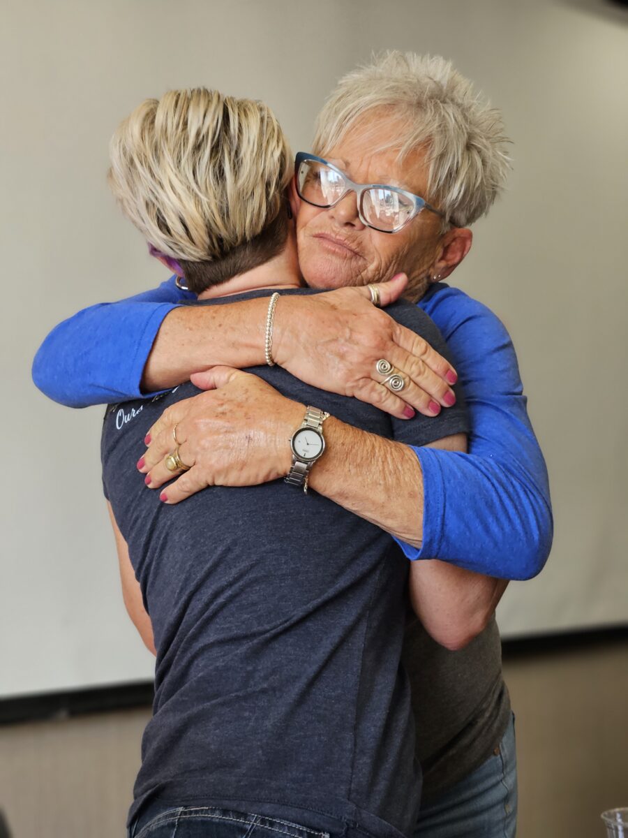 Two people, one older with white hair and glasses, and another with short, light hair, are embracing warmly. Both are wearing casual clothing, and the older person has pink-painted nails. The background is a plain, light-colored wall.