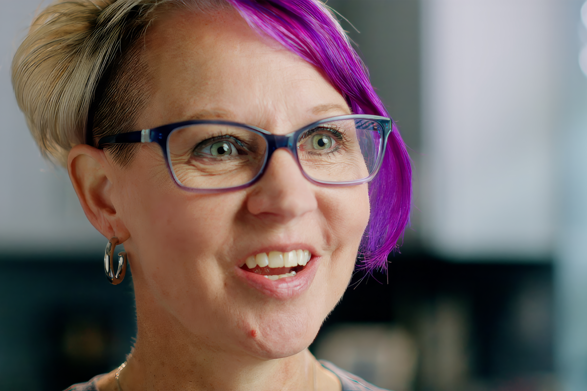 White woman with short blonde hair with a purple streak in it, and dark framed glasses.
